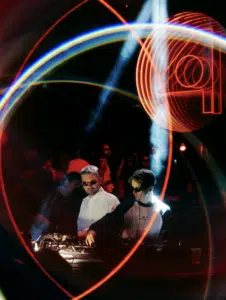 Two DJs wearing sunglasses perform at a nightclub during Amsterdam Dance Event, surrounded by colorful light trails and a neon circular design in the background.